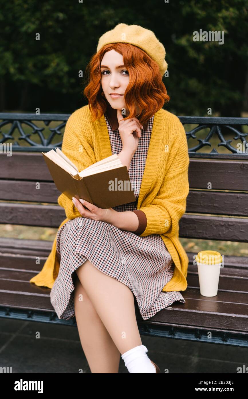 Una hermosa chica con un peinado rizado está sentado en una banca del parque y la lectura Foto de stock