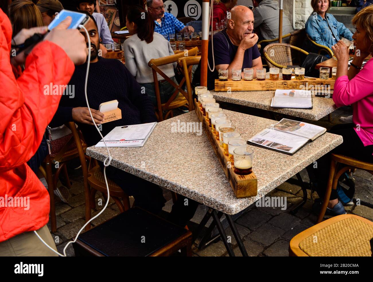 Brujas, Bélgica. Agosto De 2019. Degustación de cervezas artesanales belgas: Doce tazas dispuestas en el portavasos de madera. La gente está sentada en las mesas disfrutando Foto de stock