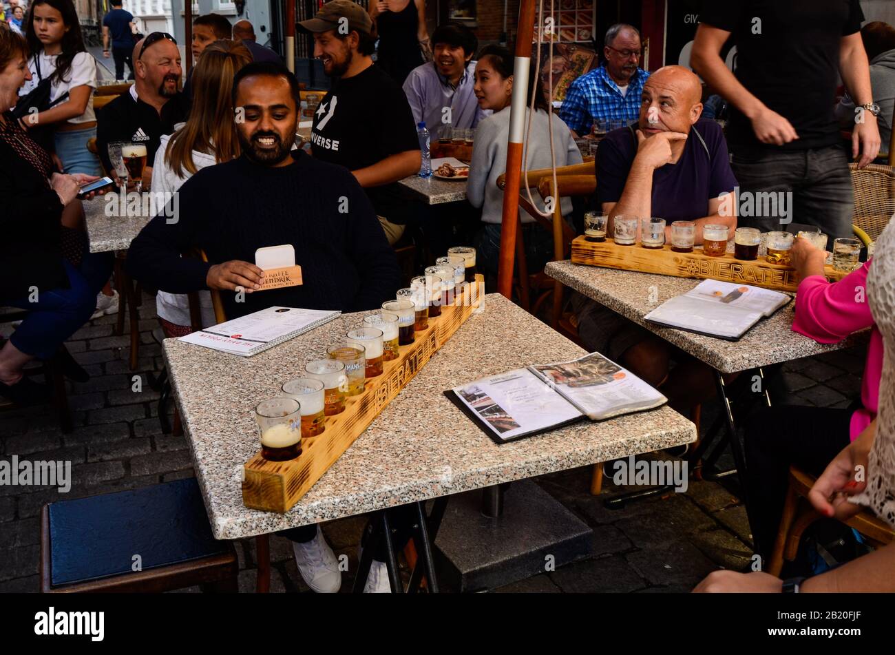 Brujas, Bélgica. Agosto De 2019. Degustación de cervezas artesanales belgas: Doce tazas dispuestas en el portavasos de madera. La gente está sentada en las mesas disfrutando Foto de stock