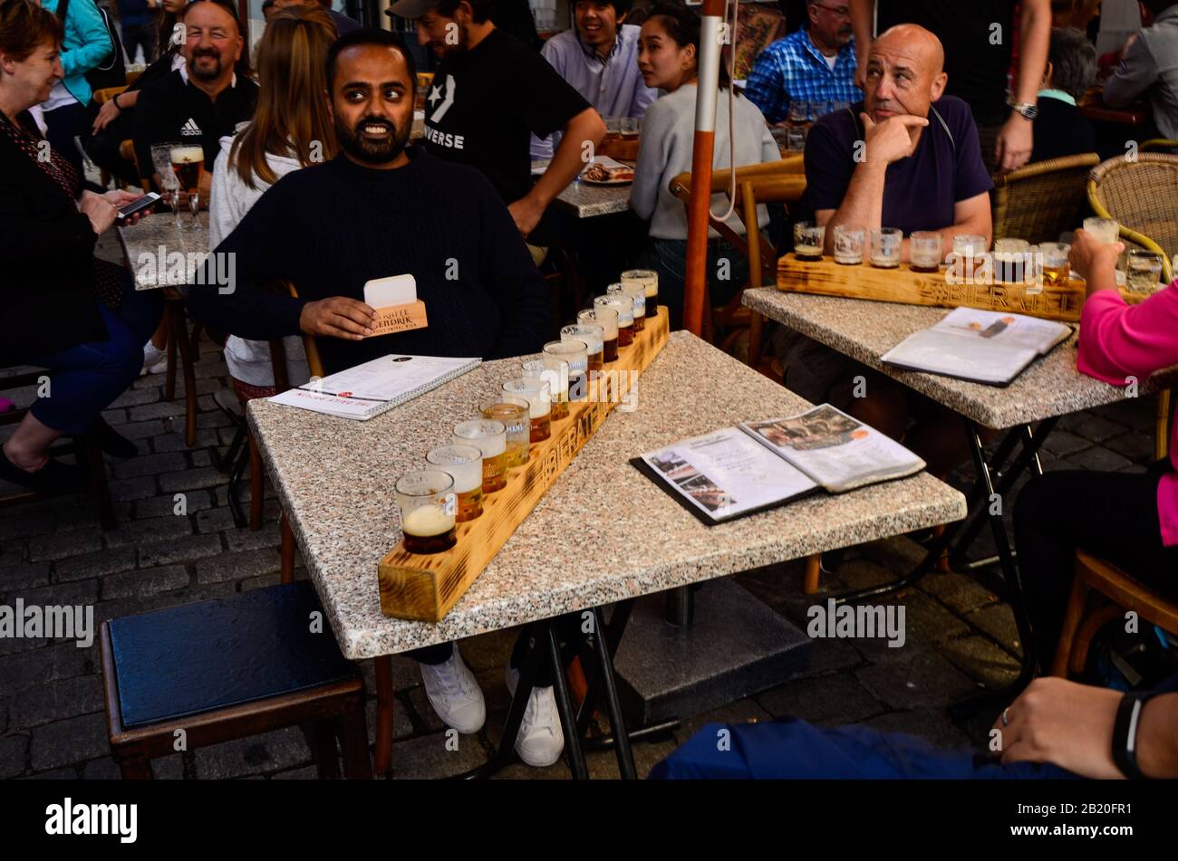 Brujas, Bélgica. Agosto De 2019. Degustación de cervezas artesanales belgas: Doce tazas dispuestas en el portavasos de madera. La gente está sentada en las mesas disfrutando Foto de stock