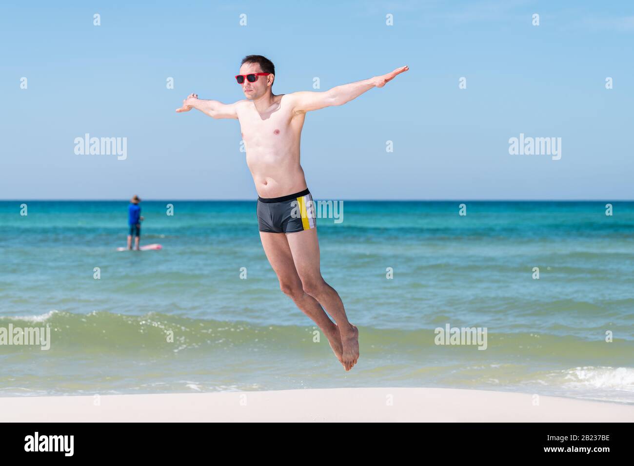 Hombre joven saltando por el agua del océano a mitad del aire con los brazos estirados en verano en Santa Rosa Beach, Florida volando con el barco en el fondo Foto de stock