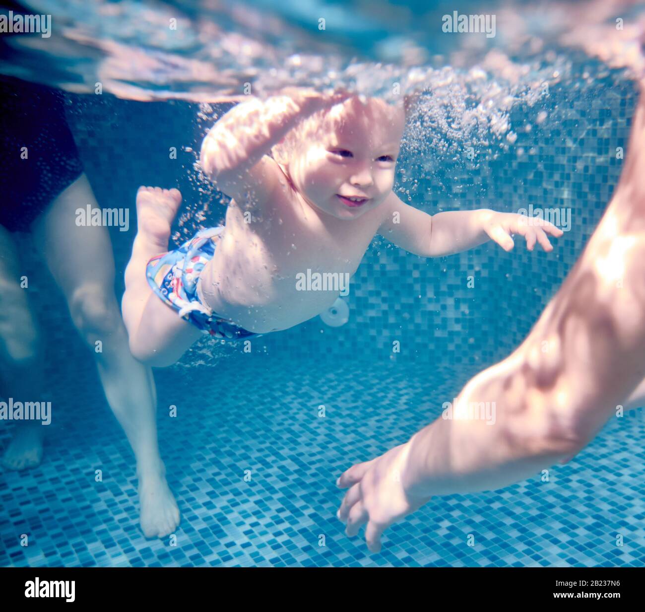 Primer plano de un lindo niño disfrutando nadando bajo el agua en la piscina. Feliz niño activo mirando a un lado y sonriendo mientras equilibra en agua azul cristalino. Concepto de la infancia, natación y actividades deportivas acuáticas. Foto de stock