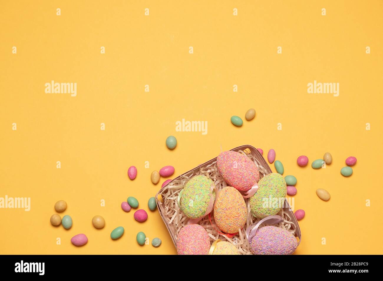 Cesta de mimbre con huevos de Pascua y deliciosos caramelos de color sobre un fondo amarillo con espacio para copias, vista superior, disposición plana. Foto horizontal Foto de stock