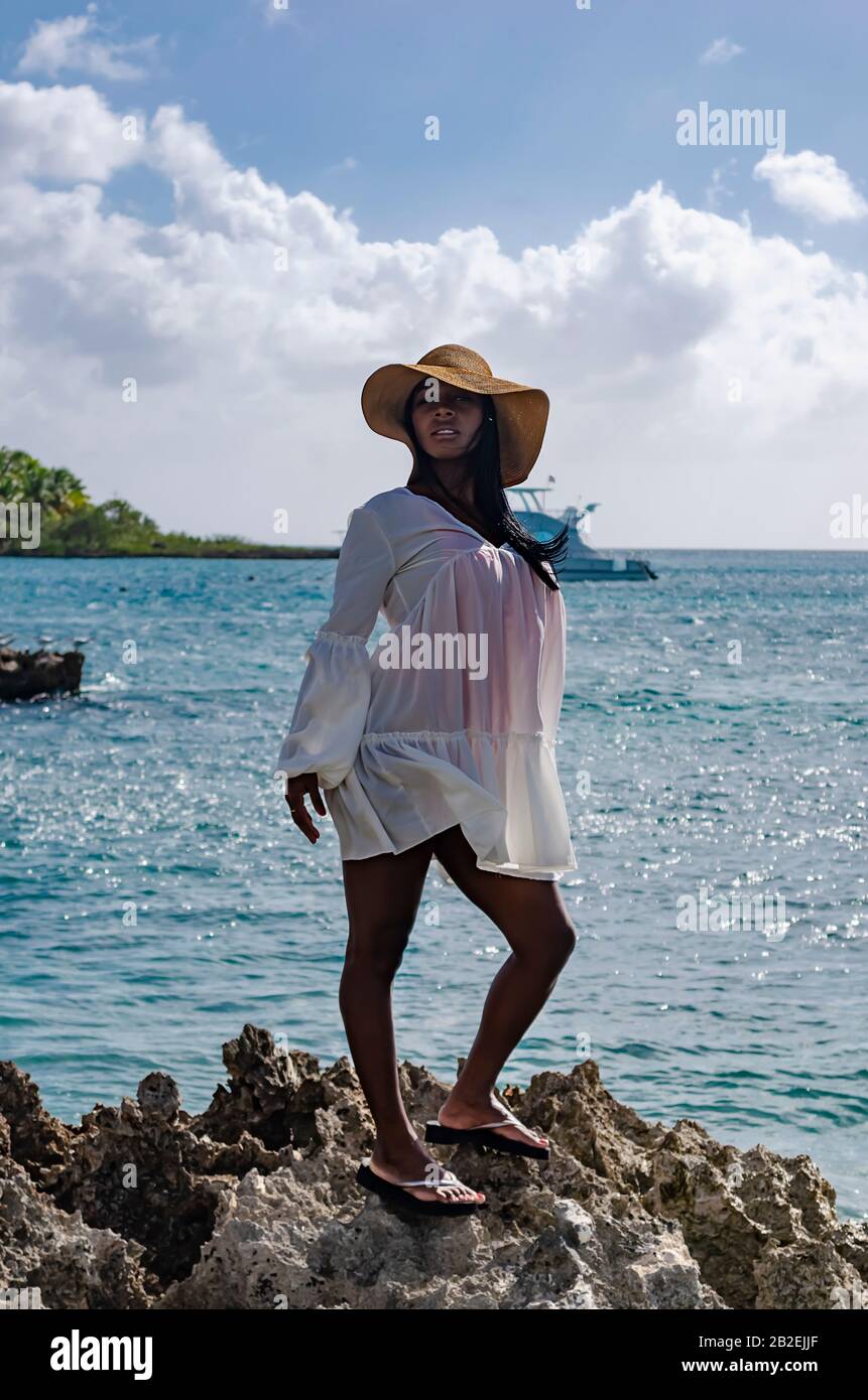 Mujer negra de 25 a 30 años de edad, de pie sobre rocas, vestida con  modelado blanco, ropa fresca de verano y tropical, con paisaje de playa y  sol s Fotografía de