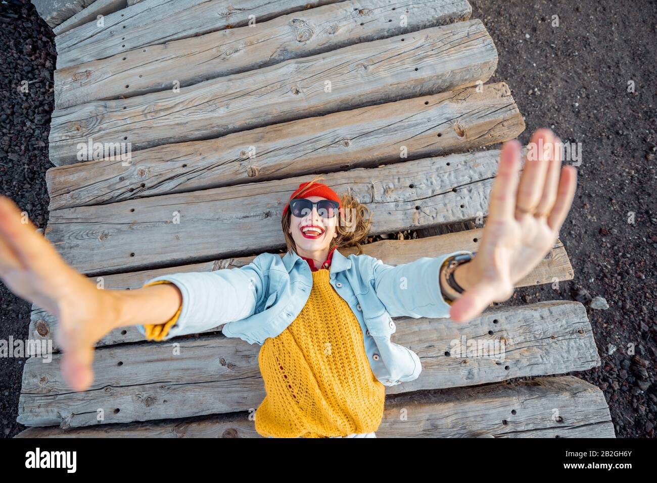 Retrato de una joven mujer con estilo vestida con ropa brillante tumbada en el camino de madera al aire libre, tirando de las manos hacia la cámara Foto de stock