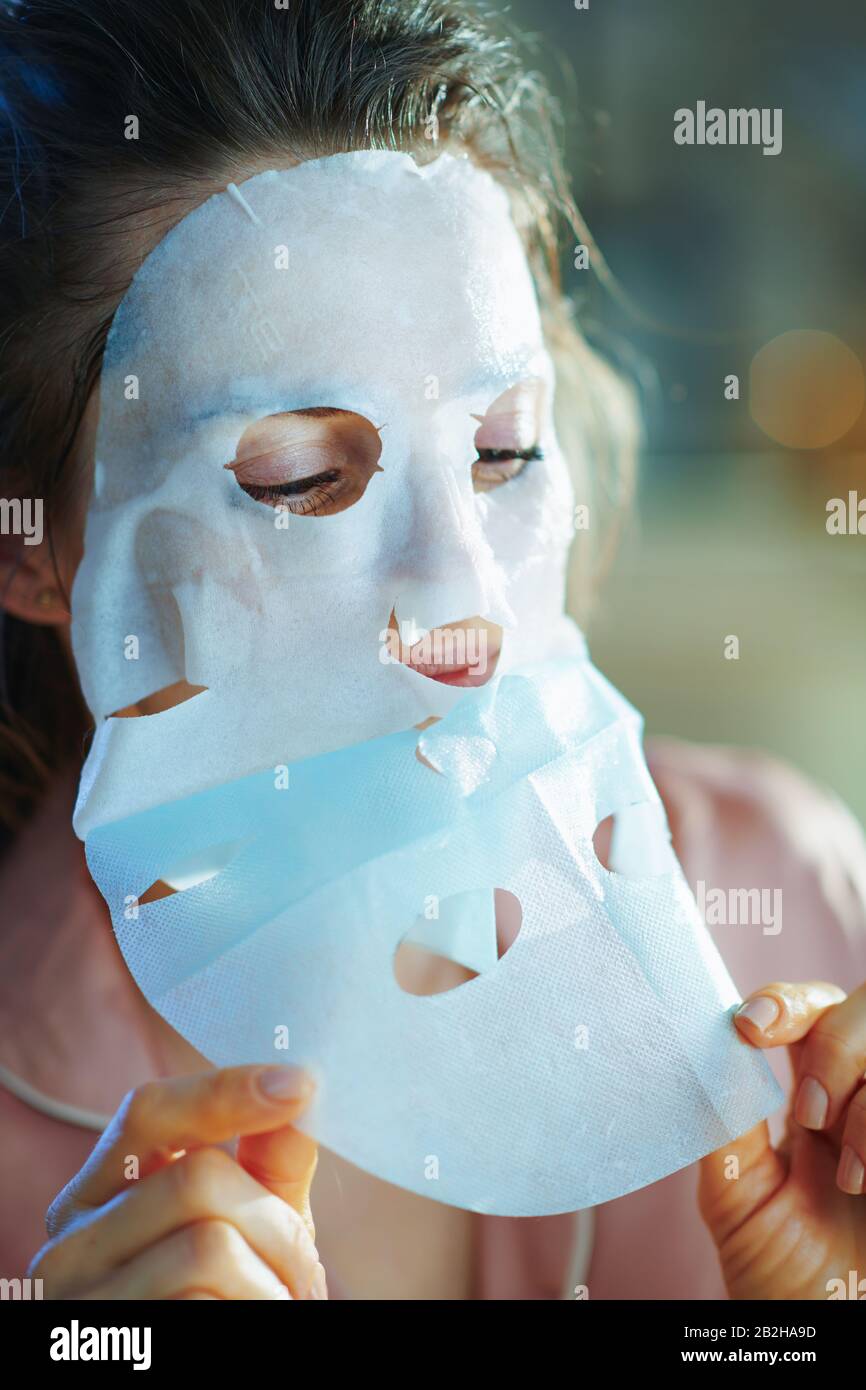 mujer joven en pijamas en el salón moderno en el soleado día de invierno aplicando una máscara facial de hoja y quitando la capa protectora. Foto de stock