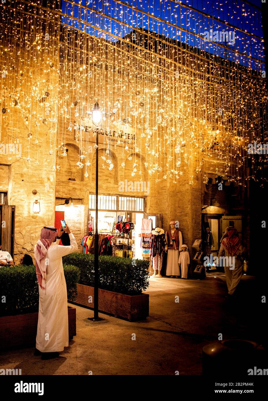 Imagen en color de un hombre de pie a lo largo de toda la longitud, vestido con ropa árabe tradicional, haciendo una foto de una pantalla de iluminación en la ciudad de night.old, Dubai. Foto de stock