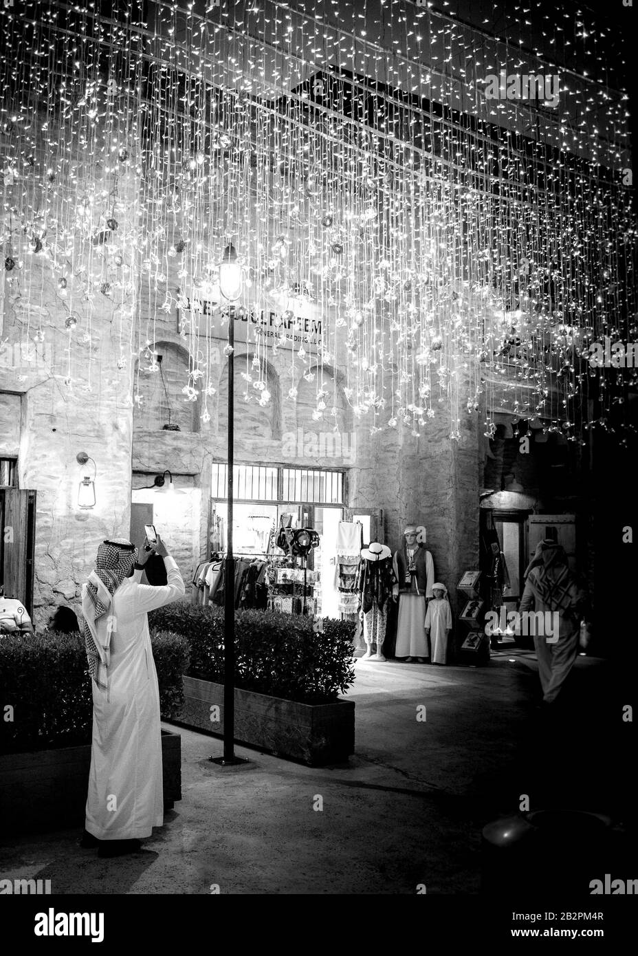 Imagen en blanco y negro de un hombre de pie de longitud completa, vestido con ropa árabe tradicional tomando una foto de una pantalla de iluminación en la ciudad de night.old, Dubai. Foto de stock