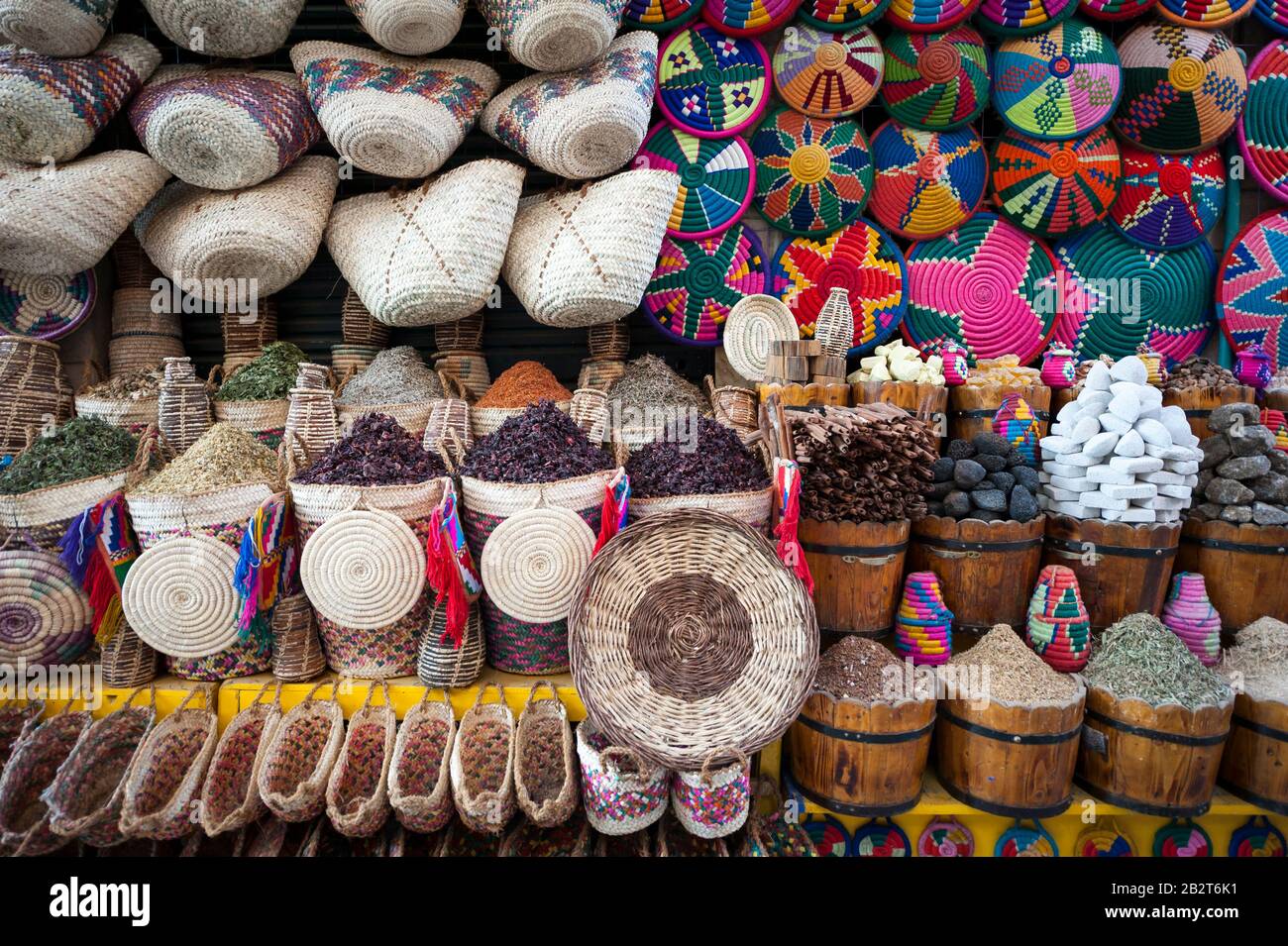Coloridas cestas y bolsas tejidas cuelgan sobre montículos de especias y hierbas en un puesto de mercado en el zoco en Asuán, Egipto Foto de stock