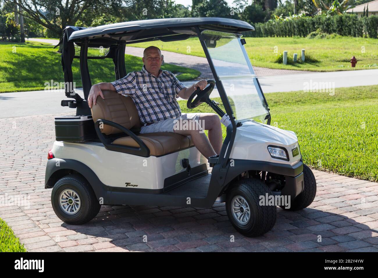 Un hombre con una camisa de cuadros y pantalones cortos se sienta en un  carrito de golf Club Car Tempo en la entrada de ladrillo de una casa de  Palm City, Florida