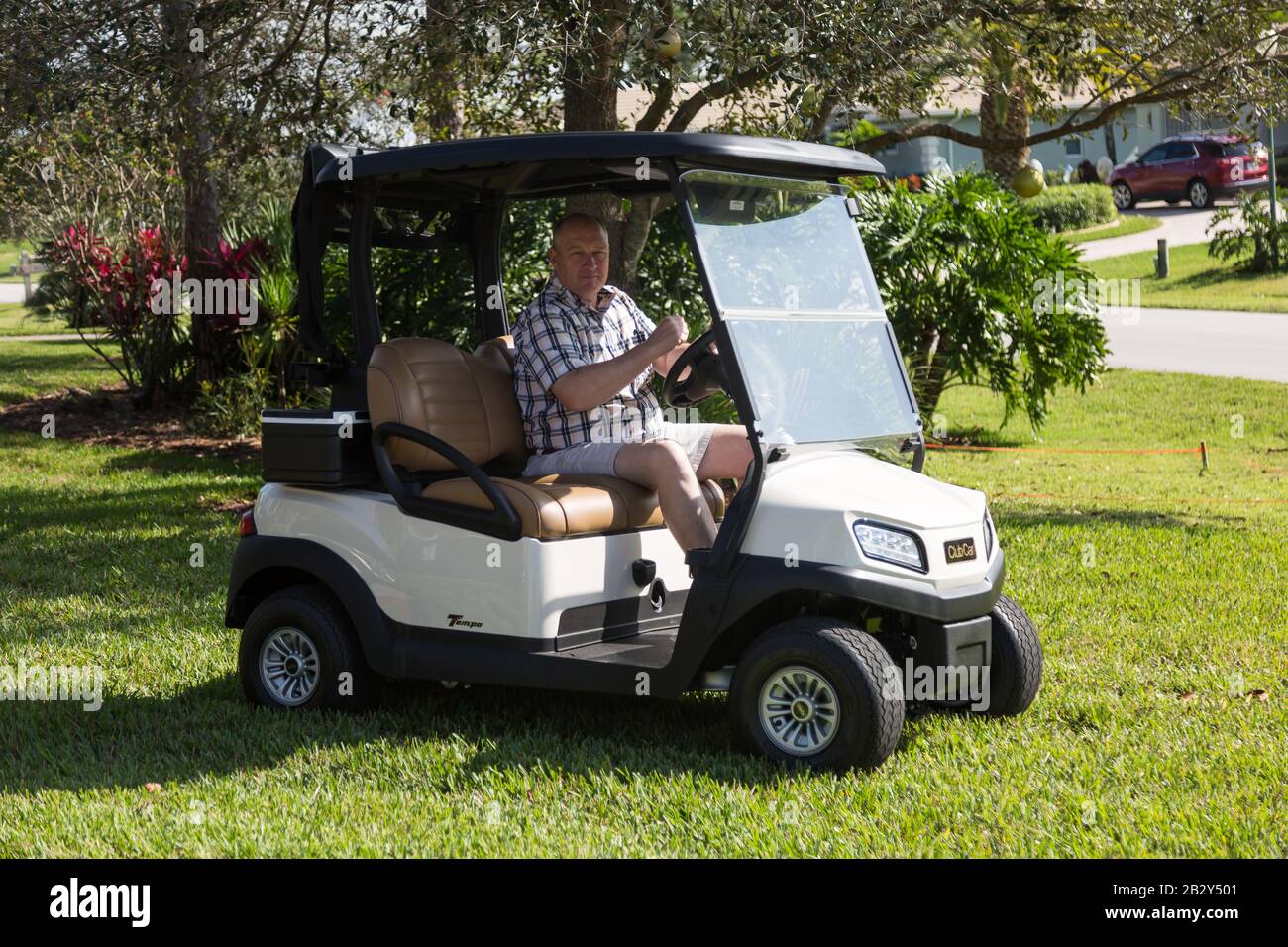 Un hombre que lleva una camisa de cuadros y pantalones cortos conduce un carrito de golf Club Car Tempo a través de la hierba de una casa en Palm City, Florida. Foto de stock