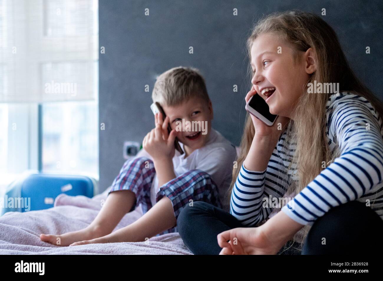 Niños exsitados el niño y la niña caucásicos hablan en los teléfonos móviles en casa. Comunicación de larga distancia, llamadas, tecnología telefónica, infancia Moderna Foto de stock