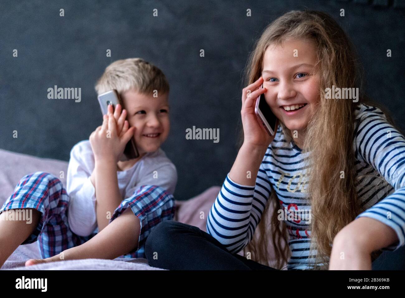 Niños exsitados el niño y la niña caucásicos hablan en los teléfonos móviles en casa. Comunicación de larga distancia, llamadas, tecnología telefónica, infancia Moderna Foto de stock