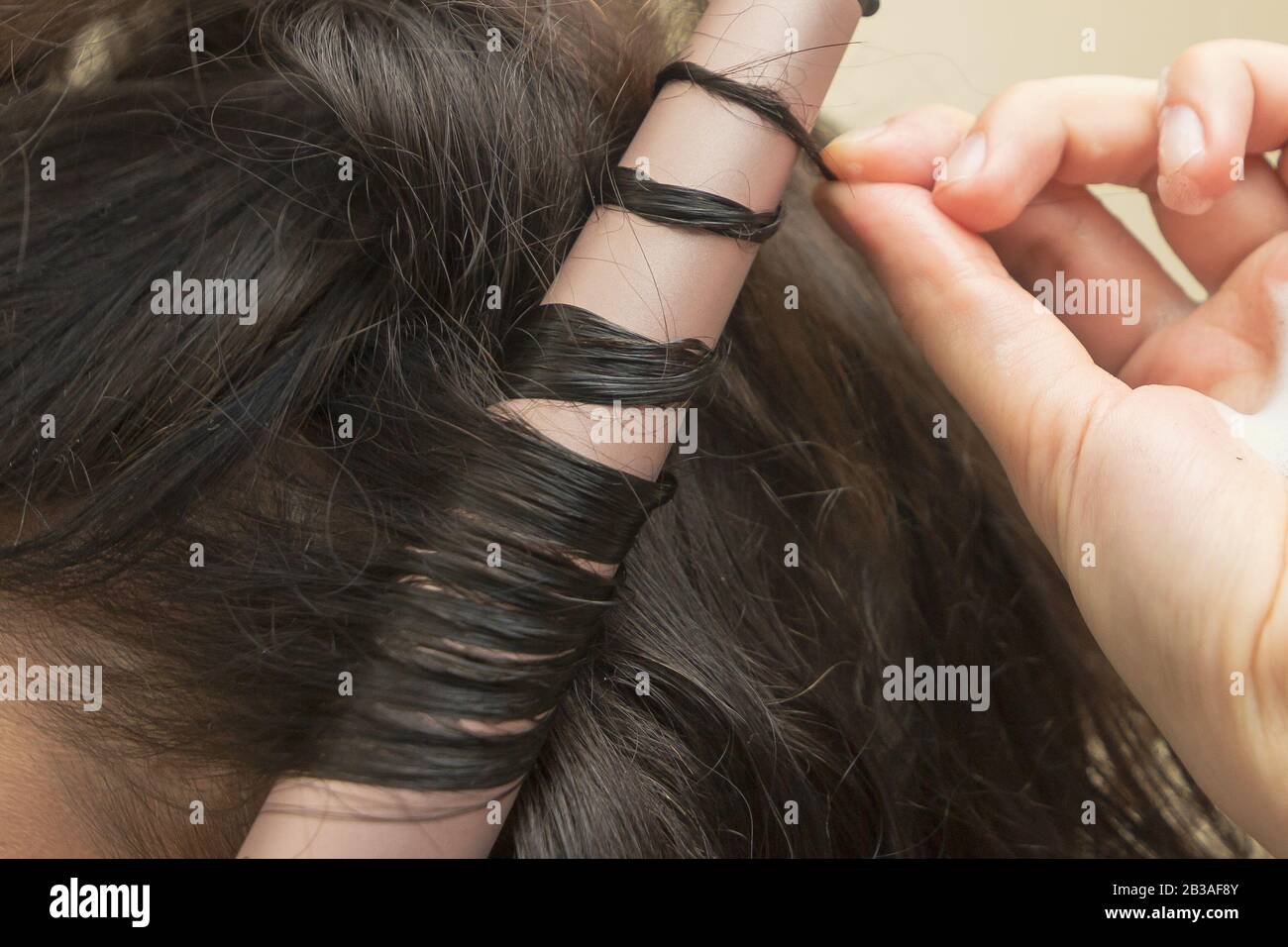 Peluquería está haciendo un pelo rizado. Un peinado de pelo con una varita ondulada cerámica. Mujer joven está moldeando su pelo recto marrón Foto de stock