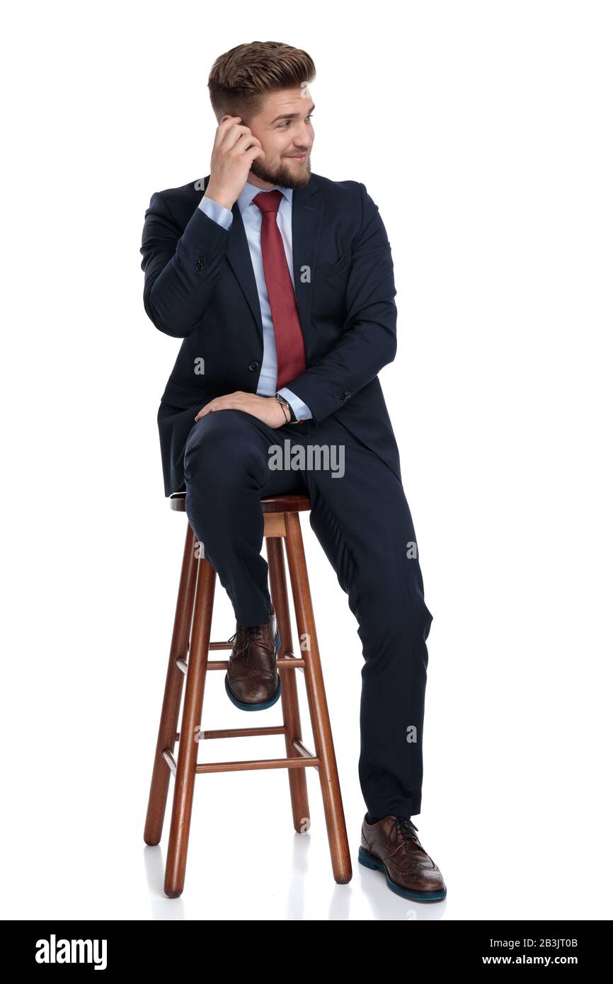 feliz joven hombre de negocios sosteniendo la mano y rascándose la cabeza, pensando y cuestionando, mirando al lado, sentado sobre fondo blanco en el estudio Foto de stock