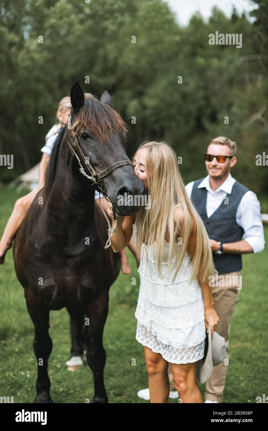 Retrato de una familia joven feliz, padres y niños, divertirse en el campo al aire libre, caminar con caballo. Los niños están montando a caballo, la madre lo es Foto de stock