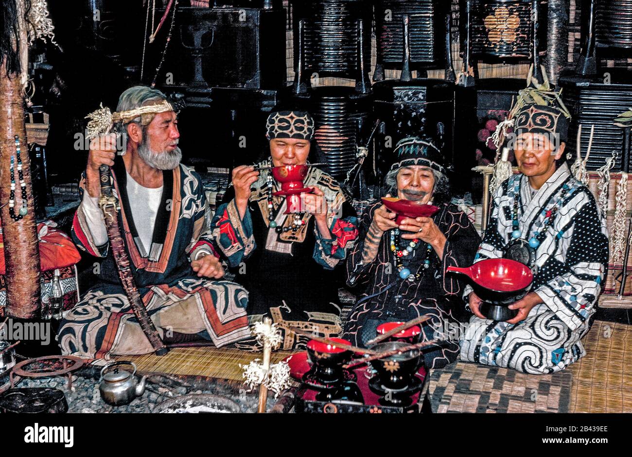 Un hombre ainu barbudo y tres mujeres posan en vestido tradicional con recuerdos ainu que venden en una casa natal que está abierta a los turistas en la isla de Hokkaido en el norte de Japón. Detrás de ellos hay barricas de laca que contienen tesoros familiares. La mujer mayor, de 84 años, tiene un tatuaje alrededor de su boca que se asemeja a un bigote. La dolorosa tradición de frotar el hollín en cortes en la piel durante la infancia de las niñas ainu para protegerse de los malos espíritus ha sido proscrita desde entonces por el gobierno japonés. Estos eran 4 de sólo 300 ainu de sangre pura que todavía viven cuando esta fotografía histórica fue tomada en 1962 Foto de stock