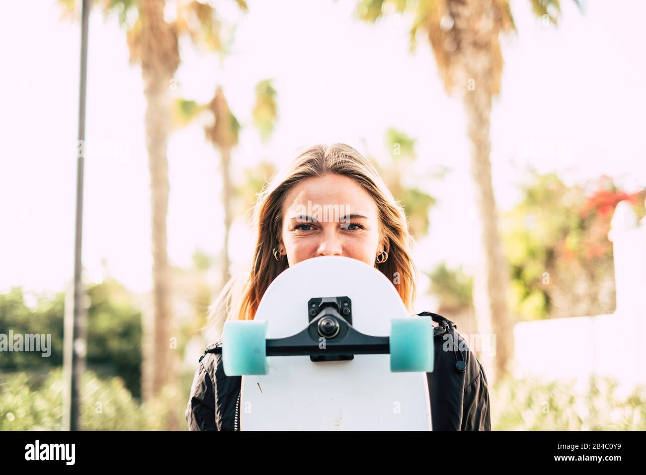 Moderno alegre hermoso retrato rubio niña con monopatín que cubre parte de la bonita cara - jóvenes milenarios personas al aire libre concepto disfrutando de estilo de vida y días soleados Foto de stock