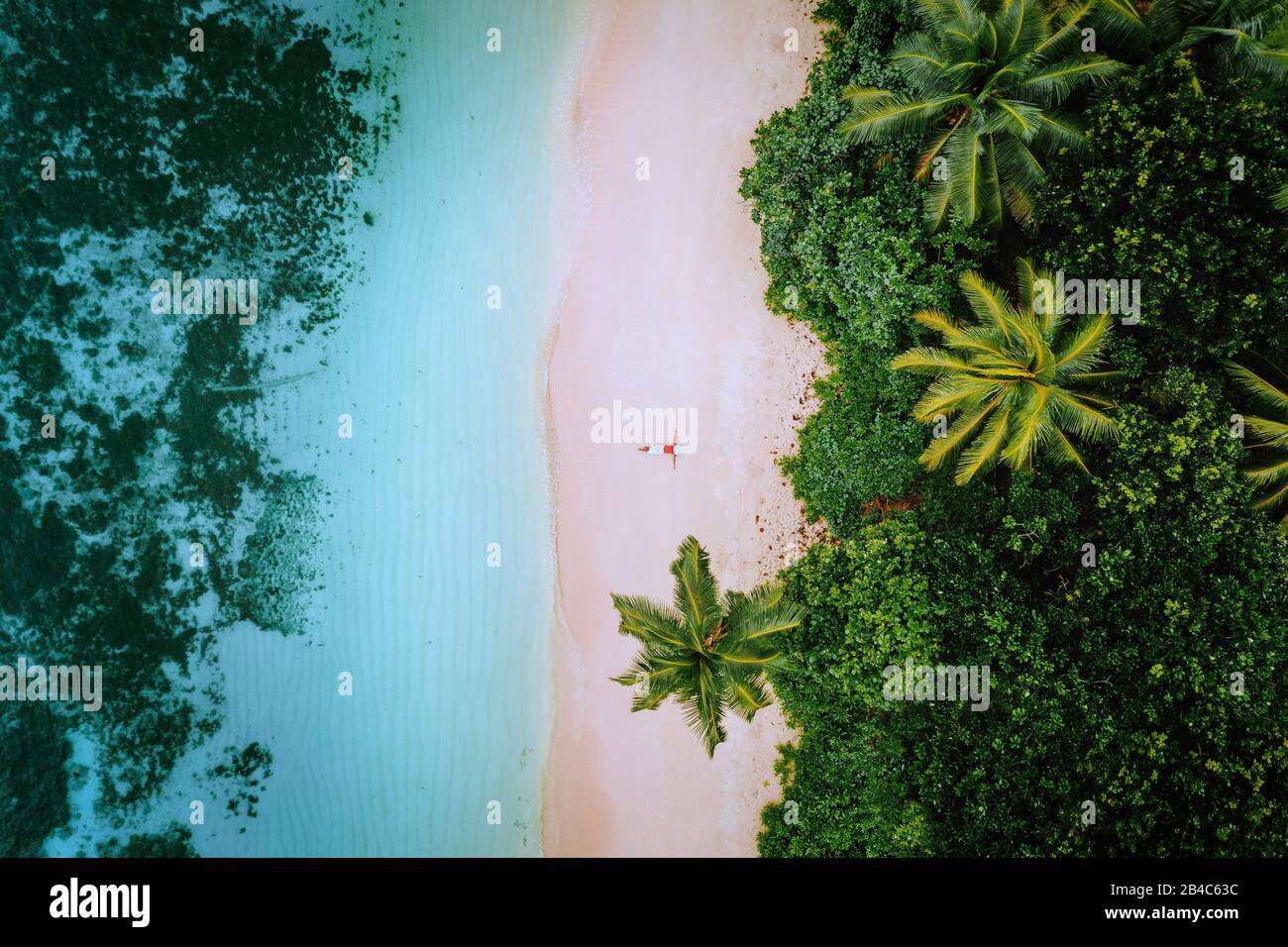 Vista aérea de una joven que se relaja en la playa de arena paradisíaca tropical rodeada de palmeras y aguas cristalinas del océano. Foto de stock