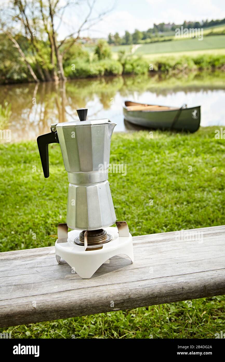 Cafetera espresso en una estufa de camping, en el fondo una canoa en el río Foto de stock