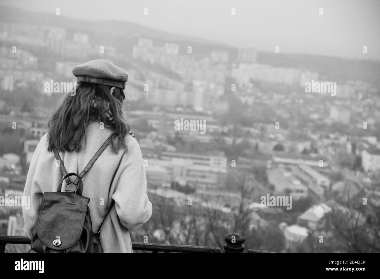 Una chica solitaria con una mochila de cuero en una gorra está de pie en una montaña alta y viendo la ciudad. Concepto de soledad Foto de stock