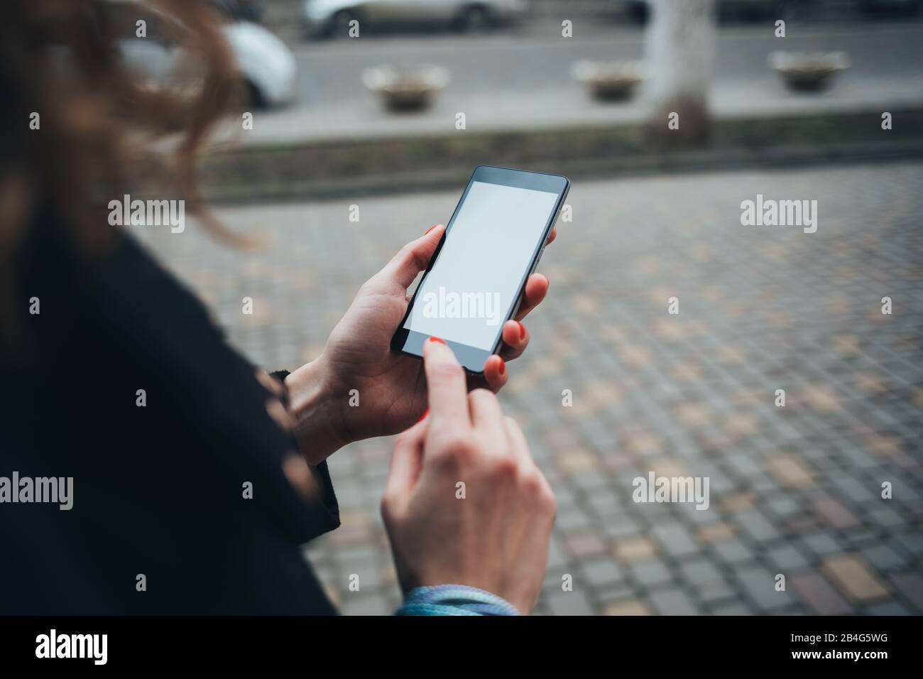Cierre las manos femeninas con manicura roja escribiendo en el teléfono inteligente con pantalla en blanco. Mujer joven utilizando el dispositivo móvil mientras camina por la calle de la ciudad en daytim Foto de stock