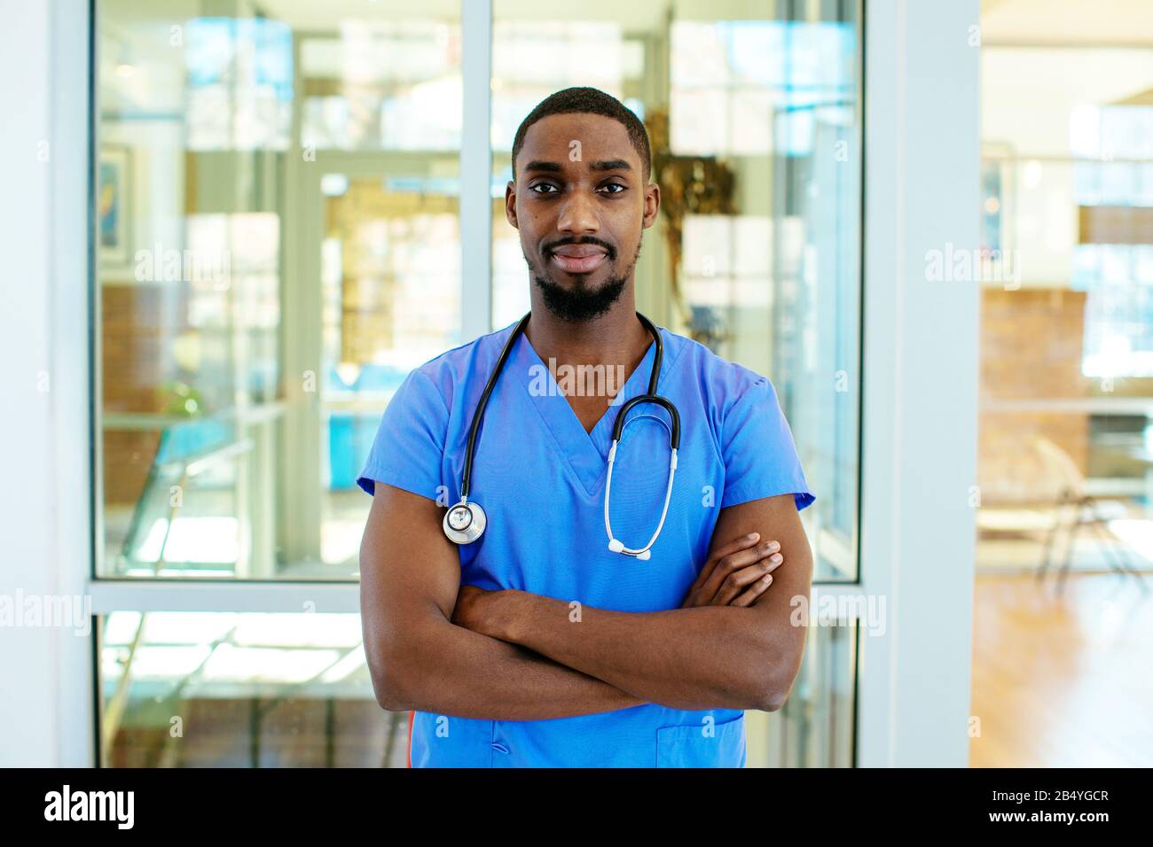 Retrato de un médico o enfermero varón joven que lleva uniforme de exfoliación azul y estetoscopio, con los brazos cruzados en el hospital Foto de stock