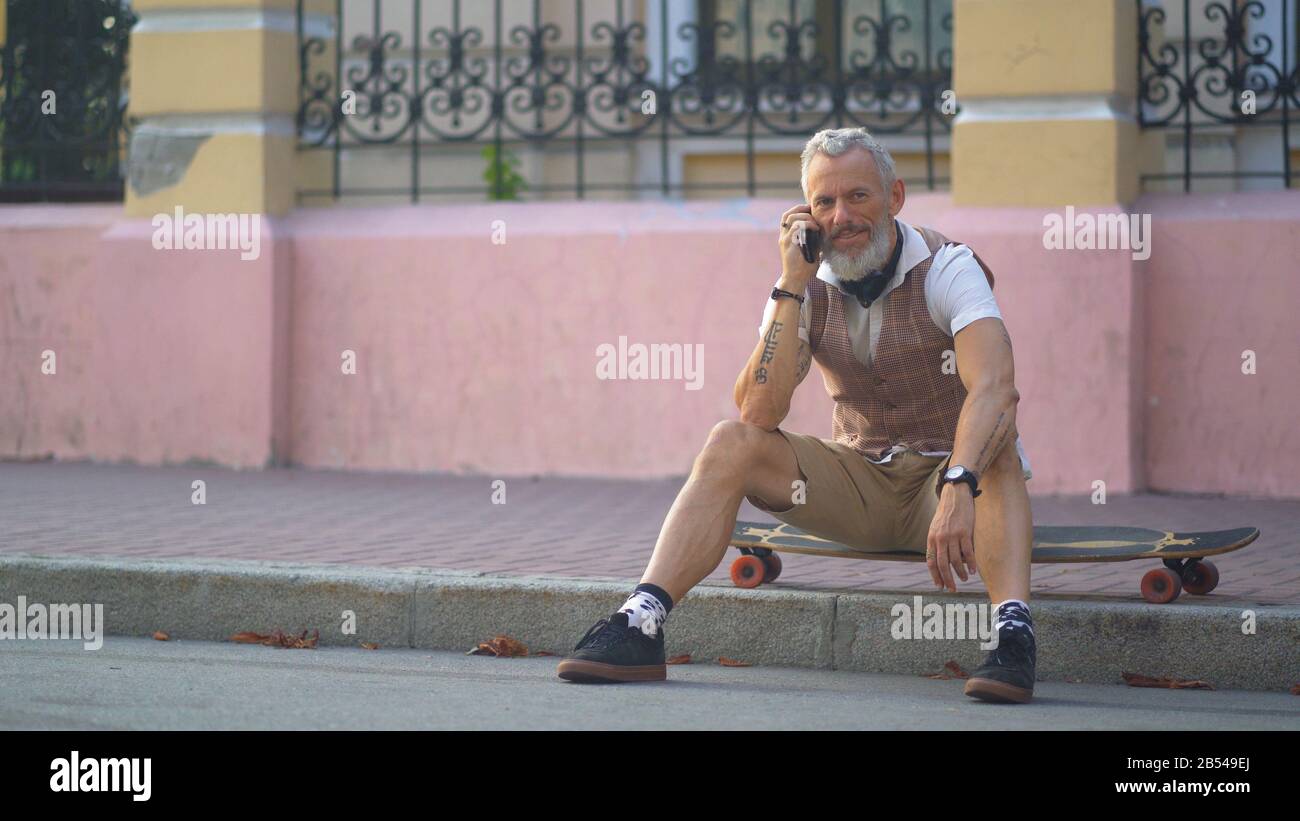El hombre de mediana edad se comunica por teléfono sentado en un monopatín en la ciudad Foto de stock
