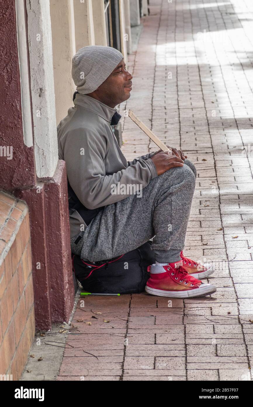 Un hombre que se sienta en la acera frente a la tienda, usando zapatos  rojos y una gorra de lana, sosteniendo un cartel de cartón en State Street,  Santa Barbara, CA Fotografía