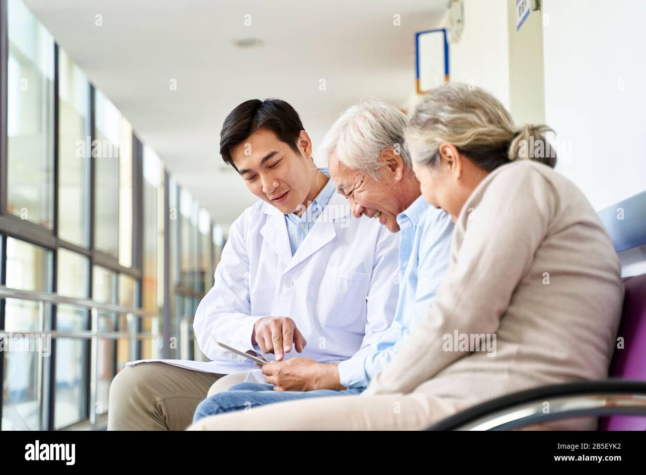 un joven médico asiático que habla sobre el resultado de la prueba y el diagnóstico con un par de pacientes mayores que utilizan tabletas digitales en el pasillo del hospital Foto de stock