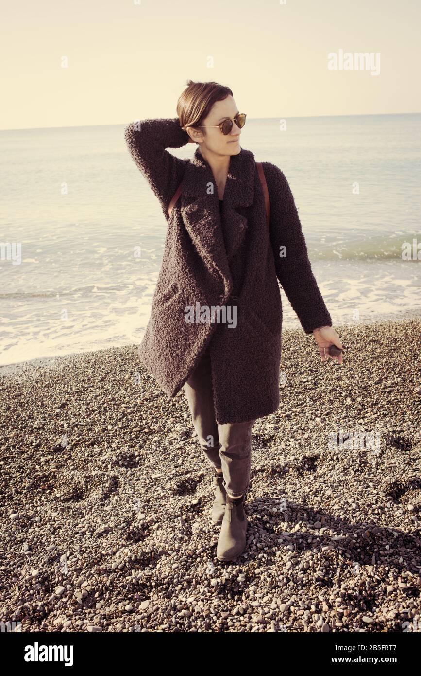 Mujer hipster modelo con gafas de sol y abrigo de invierno caminar en una playa de guijarros con el mar tranquilo vacío detrás. Efecto vintage mate. Foto de stock