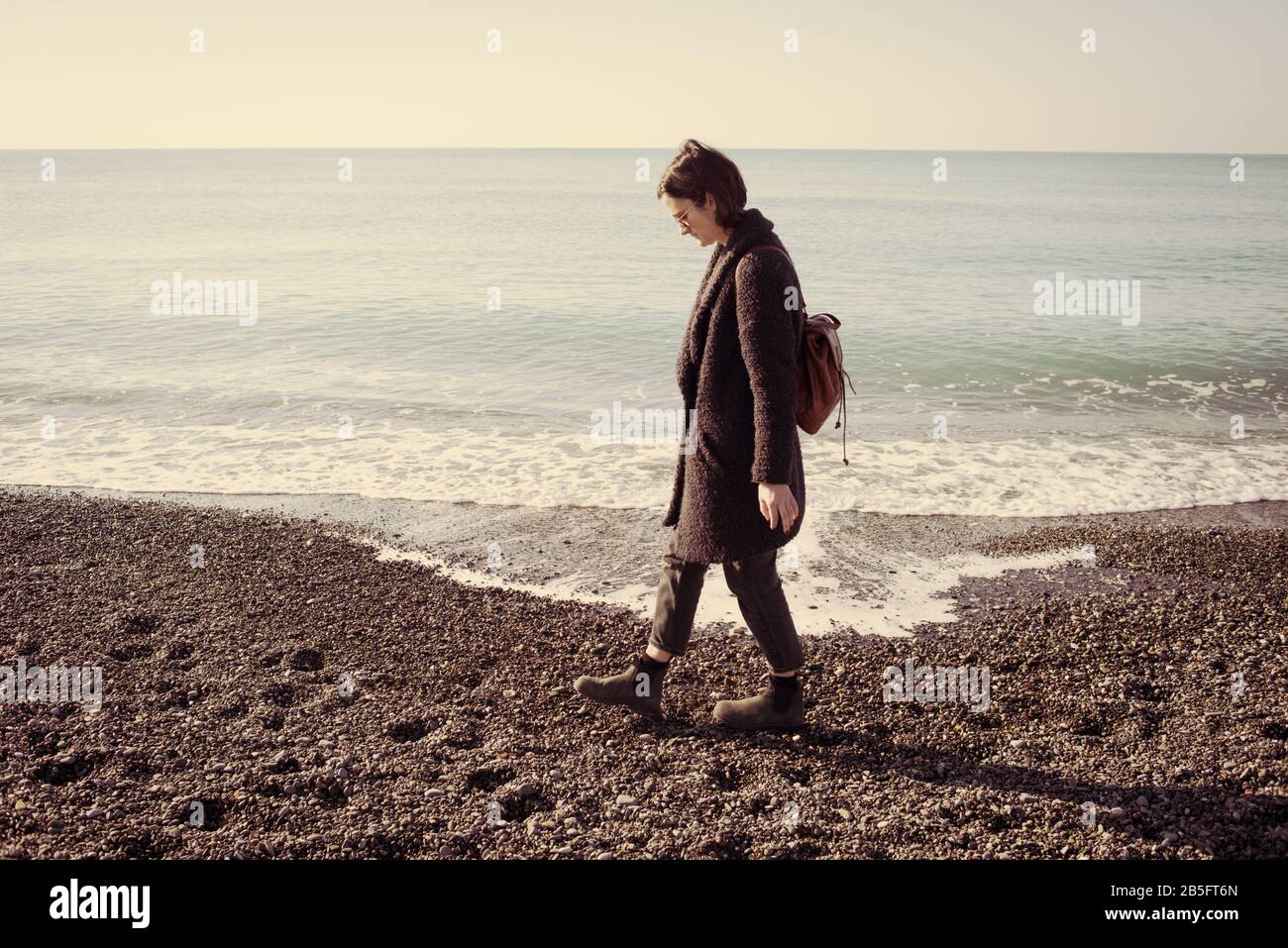 Mujer hipster modelo con gafas de sol y abrigo de invierno caminar en una playa de guijarros con el mar tranquilo vacío detrás. Efecto vintage mate. Foto de stock
