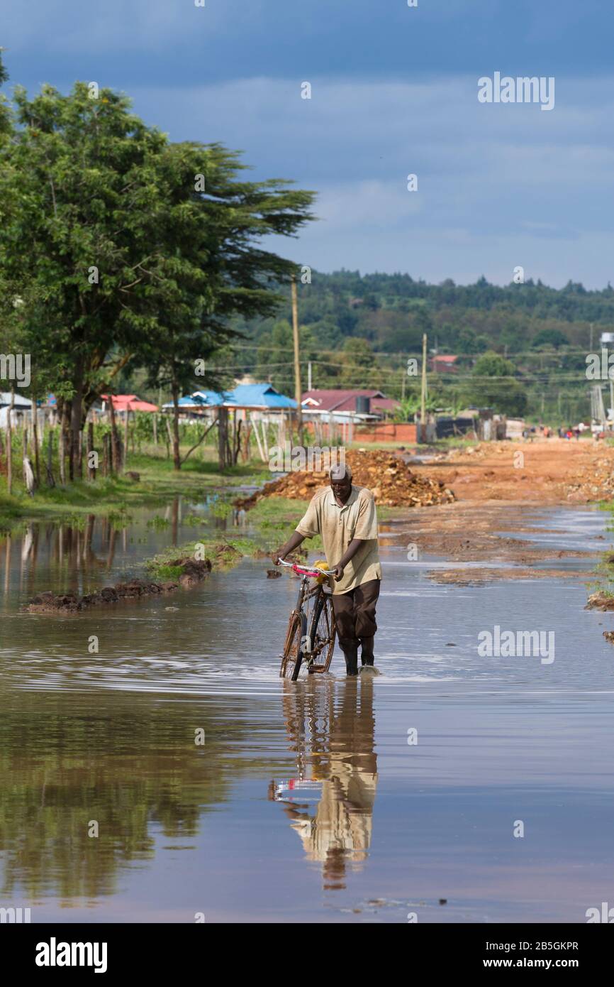 El hombre vadeando a través del agua de las inundaciones empujando una  bicicleta estilo roadster tradicional, comúnmente llamada una "Mamba Negra"  en África Oriental. Karai Rurii, Cerca De Lusiget Fotografía de stock -