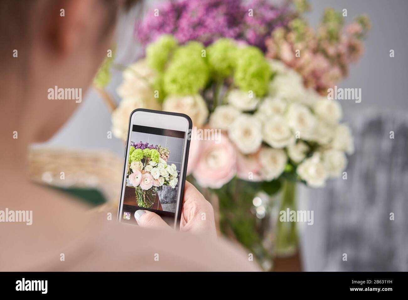 Una mujer toma una foto en su teléfono de flores. Concepto de tienda floral  . La mujer floreria crea arreglos florales en una canasta de mimbre. .  Entrega de flores Fotografía de