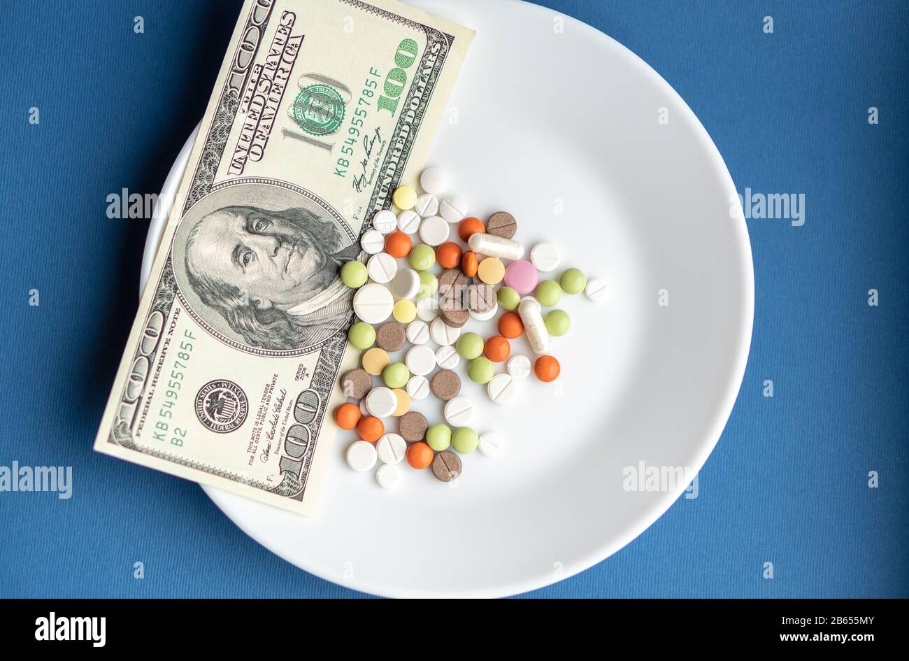 Los medicamentos cuestan dinero. Drogas y dollars.White placa con pastillas y cápsulas multicolores y un billete de 100 dólares sobre un fondo azul clásico. Pastillas Foto de stock