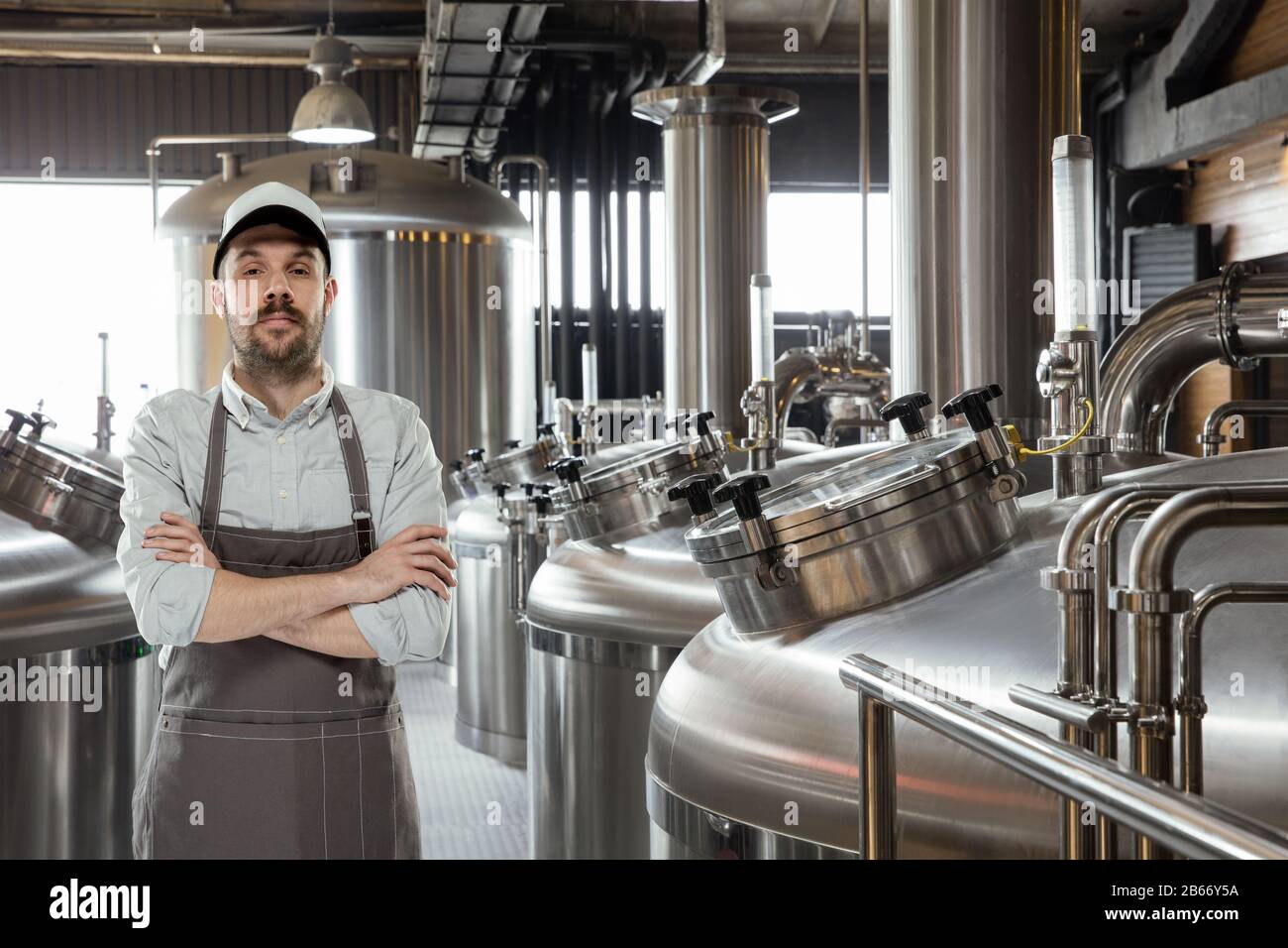 Cervecero profesional en su propia producción de alcohol artesanal.  Especialista, hombre que lleva ropa de trabajo posando con confianza con  las manos cruzadas. Concepto de empresa abierta, producto ecológico,  cervecería artesanal, fábrica