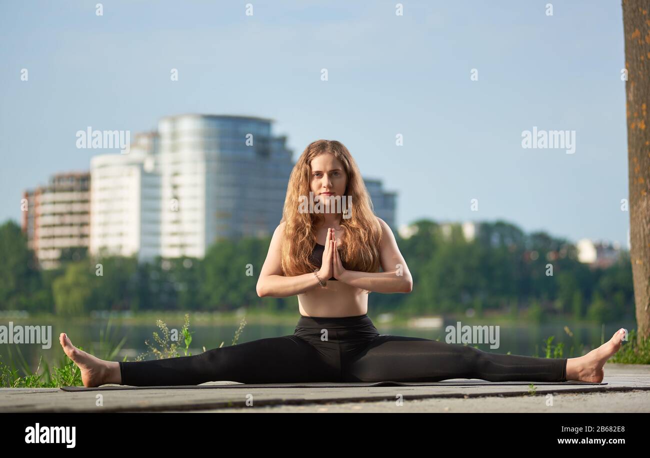 pedestal agujero Infantil Niña tierna con pelo largo practicando yoga al aire libre cerca del lago.  Chica trabajando en pantalones negros y sostén en la estera contra el fondo  borroso de los edificios de la