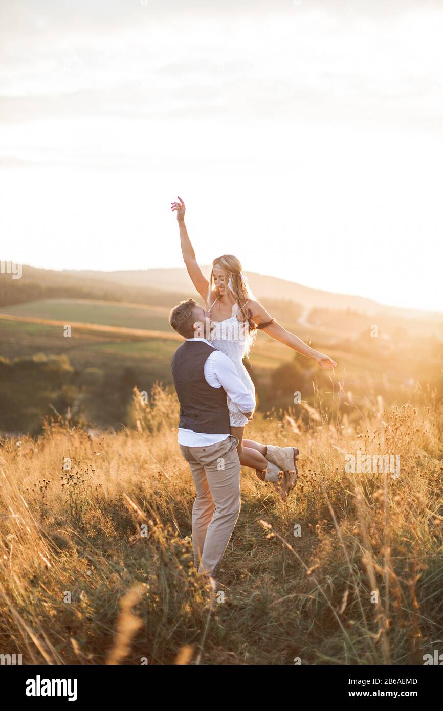 Pareja joven con ropa de estilo indie hipster boho, enamorada de caminar en  el campo, el hombre sostiene a la mujer en las manos, la mujer es feliz y  sonriente. Soleado Fotografía