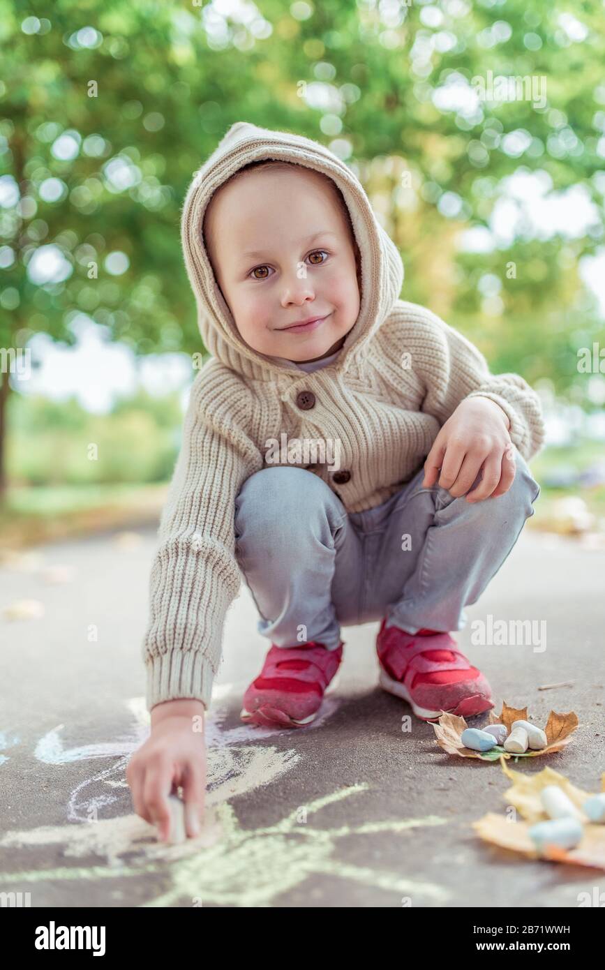El niño feliz sonríe, jumper beige con capucha, día de en verano en la ciudad, dibuja el sol sobre asfalto con tizas coloridas. Concepto creativo idea y Fotografía de stock -