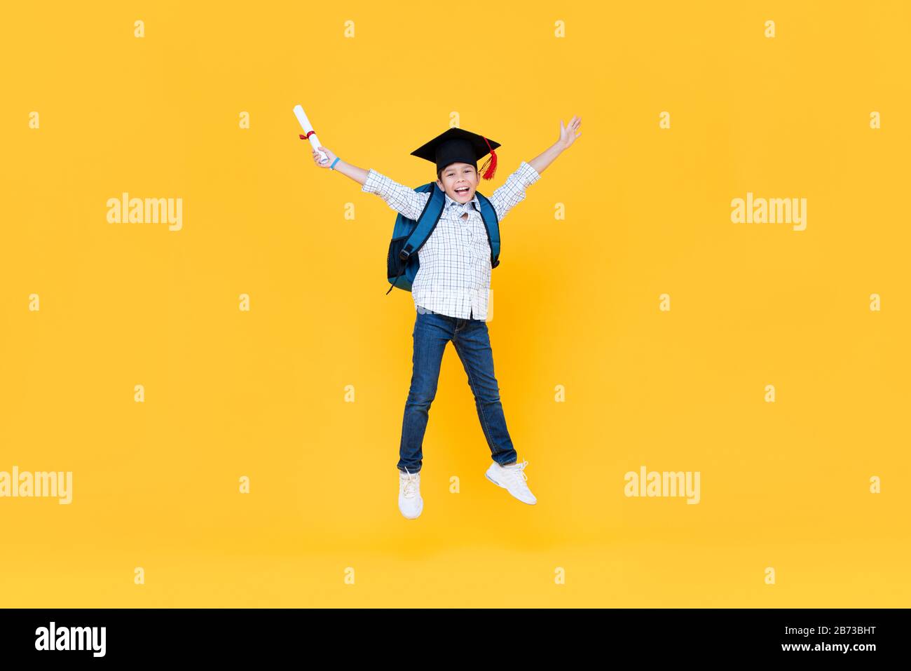 Guapo escolar con gorra de graduado sonriendo y saltando sobre fondo amarillo para el concepto de la educatina Foto de stock