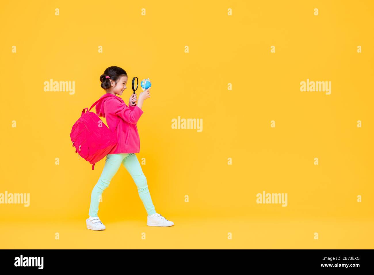 Sonriente linda bonita chica con pelo pigtail en rosa chaqueta y mochila mirando a través de una lupa en amarillo aislado fondo de estudio Foto de stock