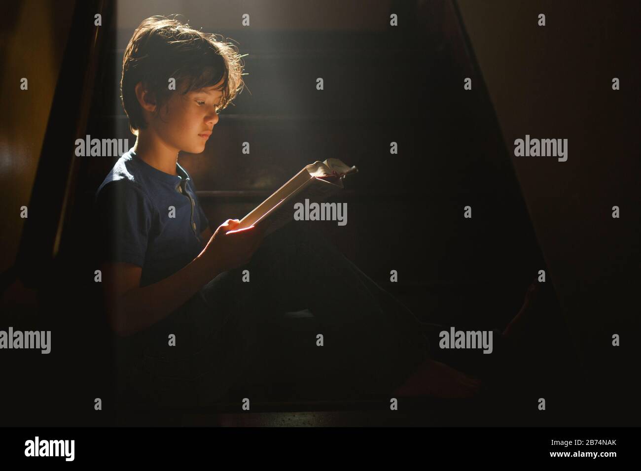 Un niño se sienta en una escalera oscura en un rayo de luz brillante de lectura Foto de stock
