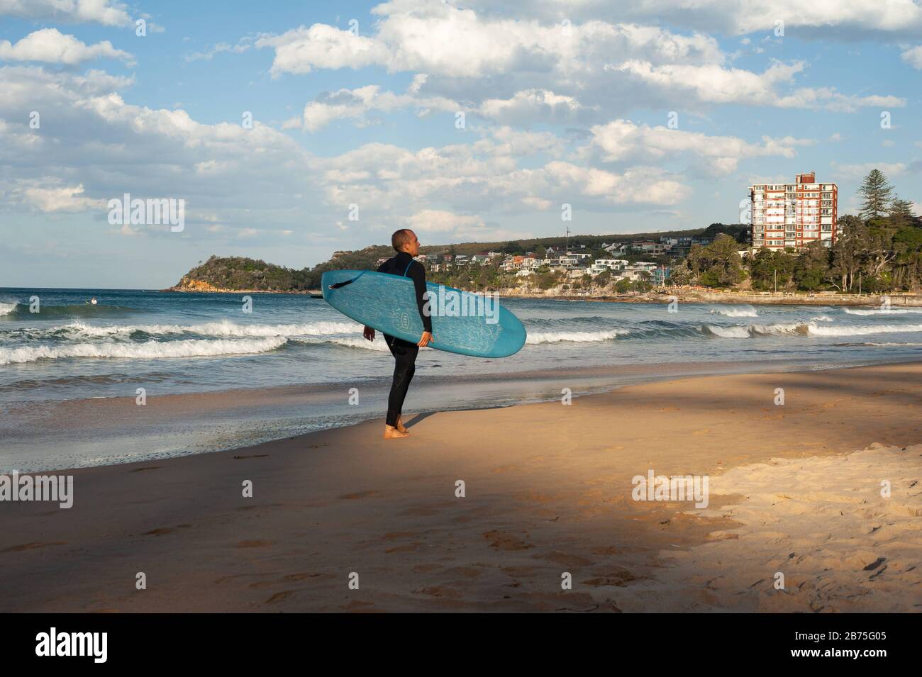 10.05.2018, Sydney, Nueva Gales del Sur, Australia - un hombre camina a lo largo de la playa de Manly con su tabla de surf bajo su brazo mientras otros surfistas están esperando en el fondo para la ola derecha. [traducción automática] Foto de stock