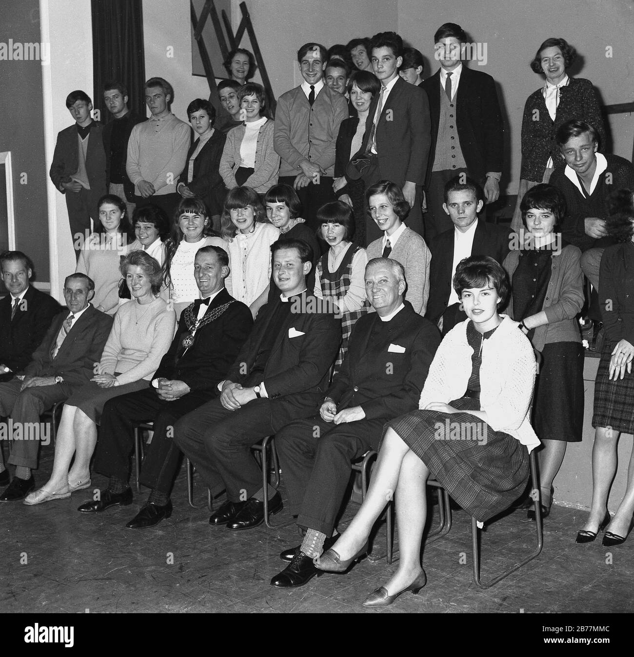 1960, histórico, grupo de jóvenes excitados en un salón de pueblo con una imagen de invitados distinguidos incluyendo el alcalde y los sacerdotes locales, Inglaterra, Reino Unido. Foto de stock