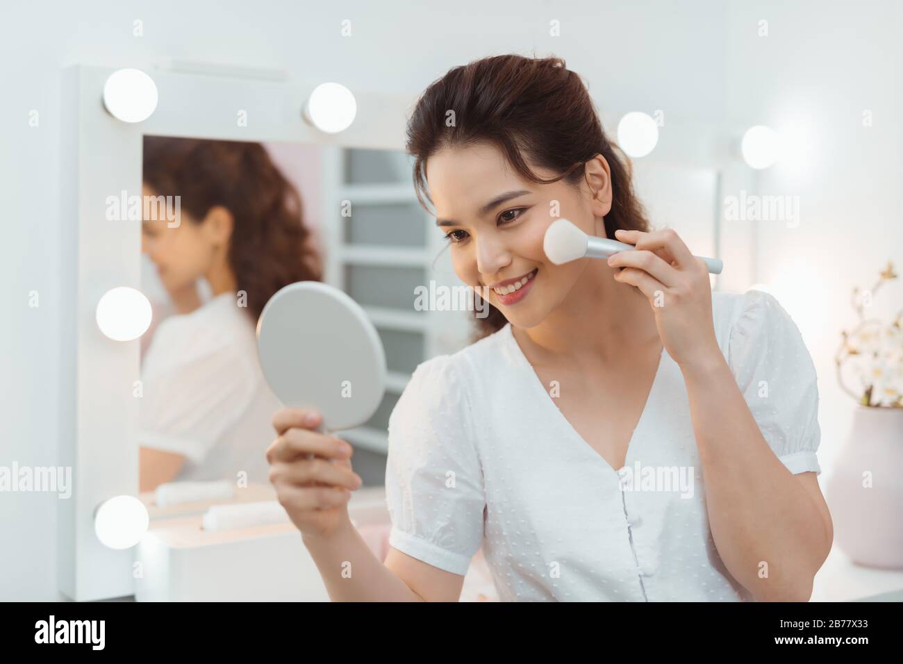 Modelo de belleza niña aplicando maquillaje y sonriendo. Hermosa mujer joven mirando en el espejo y aplicando cosméticos con un gran cepillo. Foto de stock