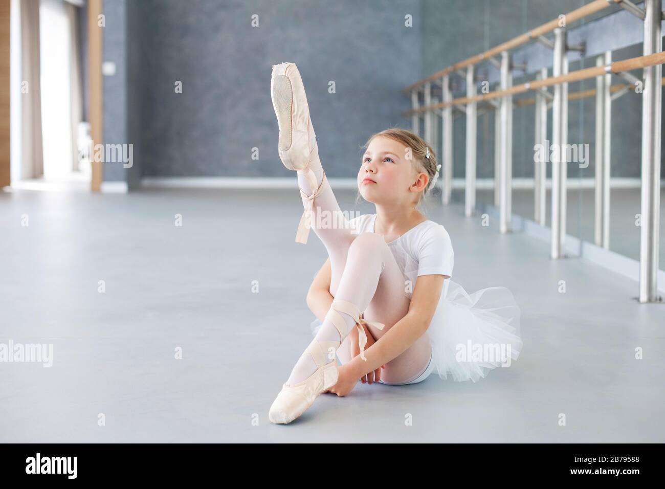 La bailarina pequeña está intentando en zapatos de la punta en la clase del ballet. Linda niña está sentada en el suelo bajo barre Foto de stock