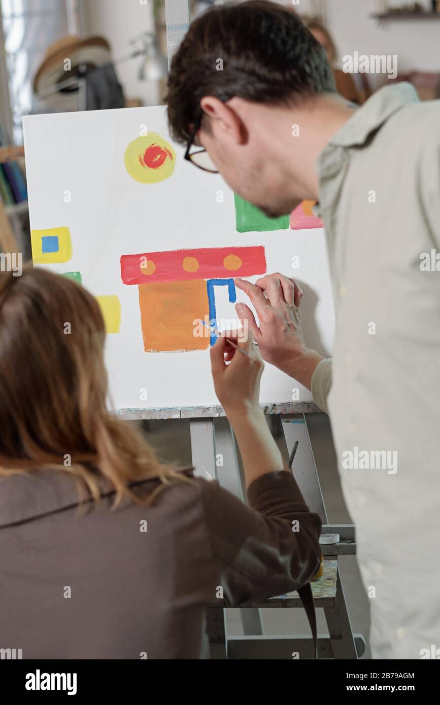 Profesor de la escuela de artes que consulta a uno de sus estudiantes en la lección mientras que está parado pintando y apuntando a la línea azul Foto de stock
