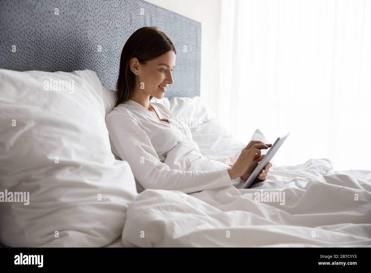 Mujer sonriente tumbada en la cama bajo el edredón, usando una tableta digital. Foto de stock
