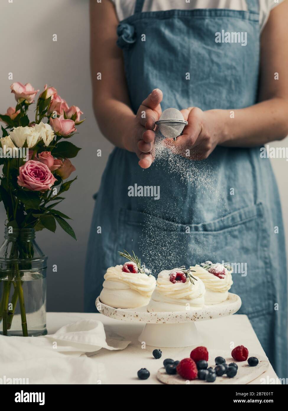 Mujer en un delantal espolvorea azúcar glaseado a través de un tamiz en mini pasteles Pavlova con bayas frescas. Composición vertical con mesa de mármol blanco an Foto de stock