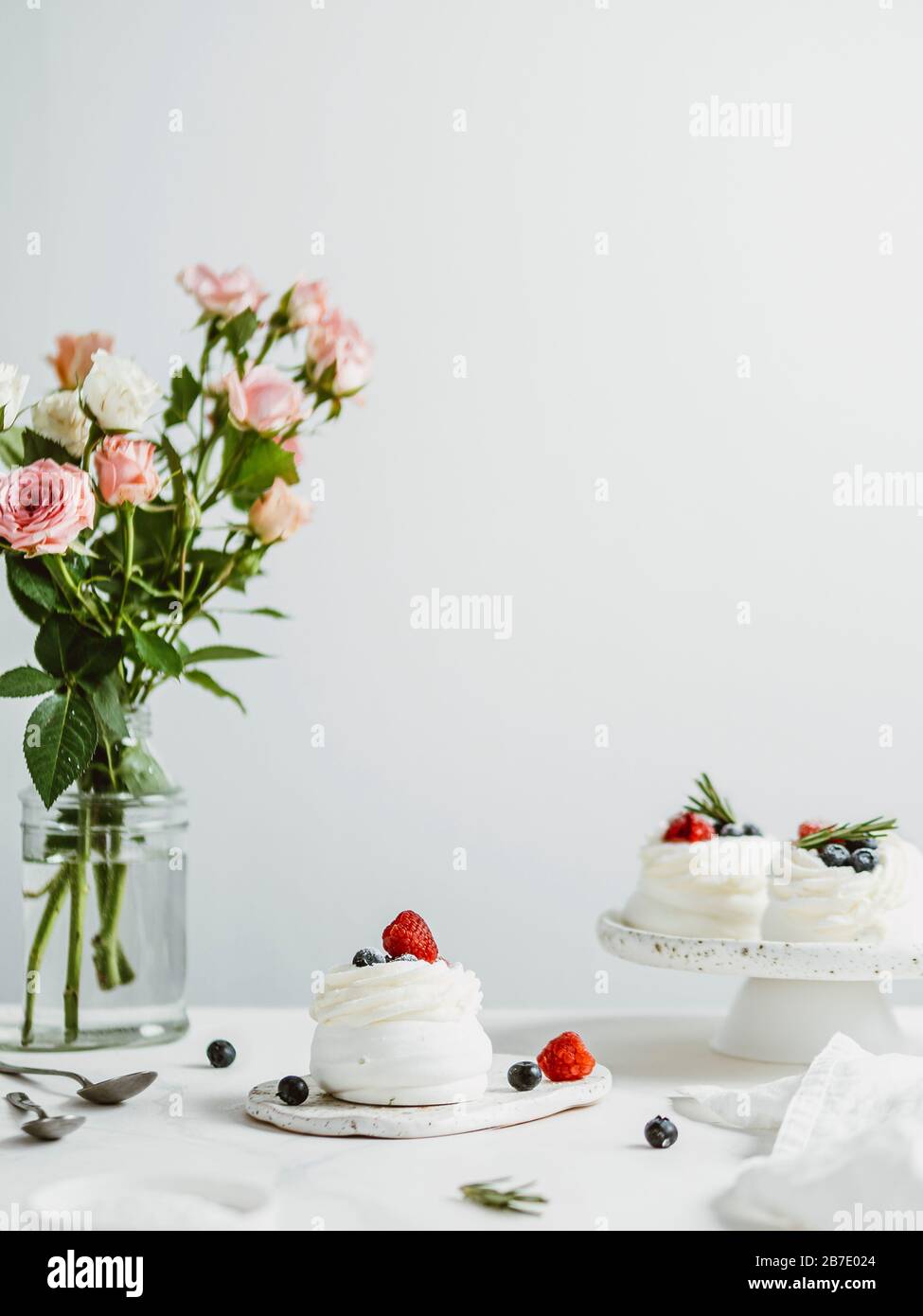 Mini pasteles Pavlova con bayas frescas y romero sobre mesa de mármol blanco con flores en botella de jarrón de cristal y fondo blanco de pared. COM. Vertical Foto de stock